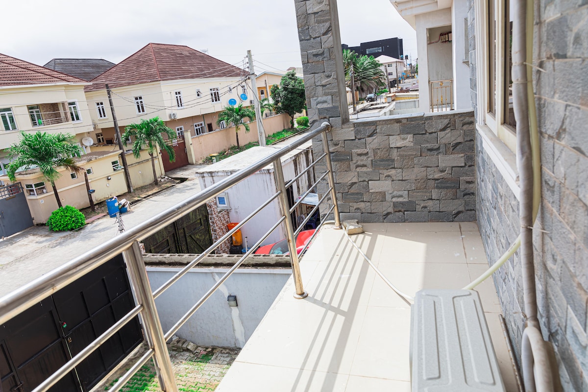 A balcony features a sleek railing, offering an unobstructed view of the surrounding area. The stone exterior of the building is visible, while the tiled floor provides a clean and spacious appearance. Nearby structures and greenery can be seen in the background.