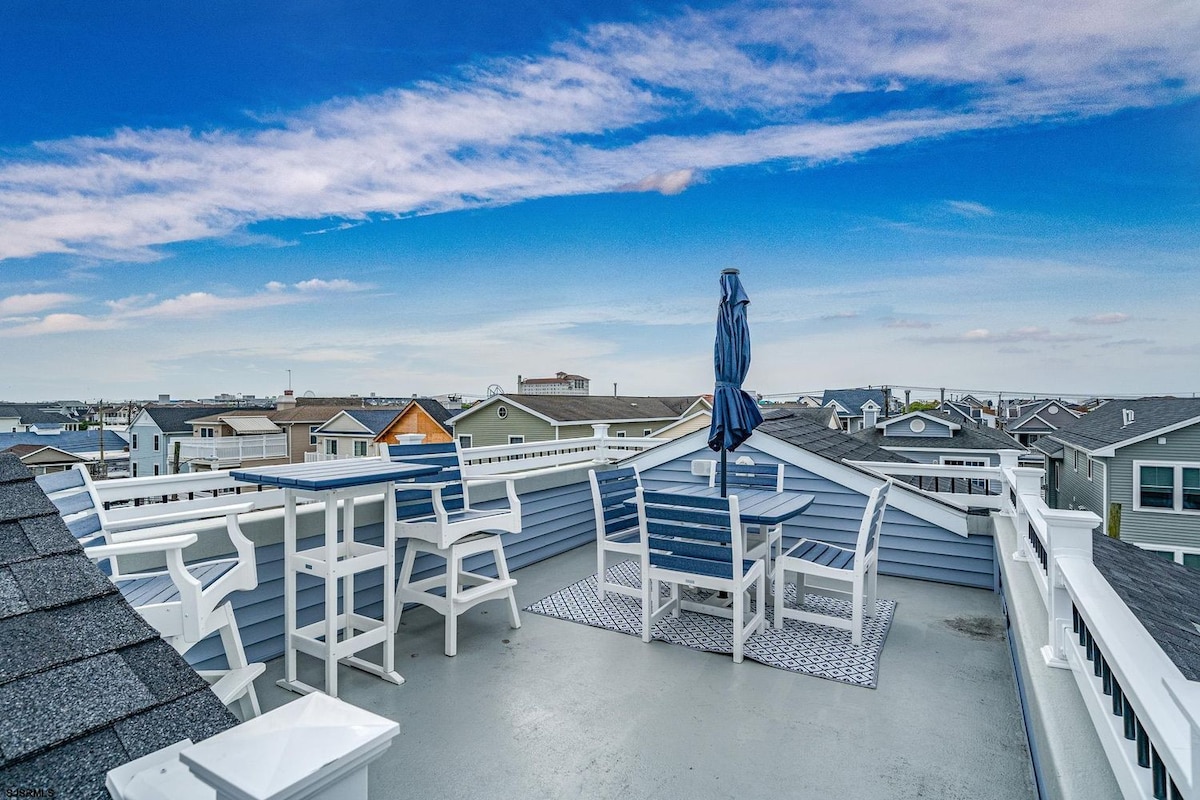 An expansive roof deck is presented, featuring a stylish dining table and several white lounge chairs. A blue umbrella provides shade, complementing the clear blue sky. The surrounding rooftops of neighboring homes are visible, showcasing a serene coastal atmosphere.