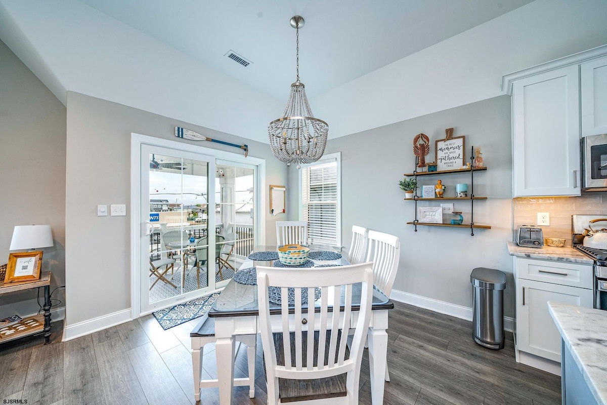 An airy dining area features a round table surrounded by white chairs, illuminated by a pendant light. Glass doors lead to a balcony, providing natural light. Multicolored decor accents the light gray walls, and open shelving displays decorative items and kitchen essentials.