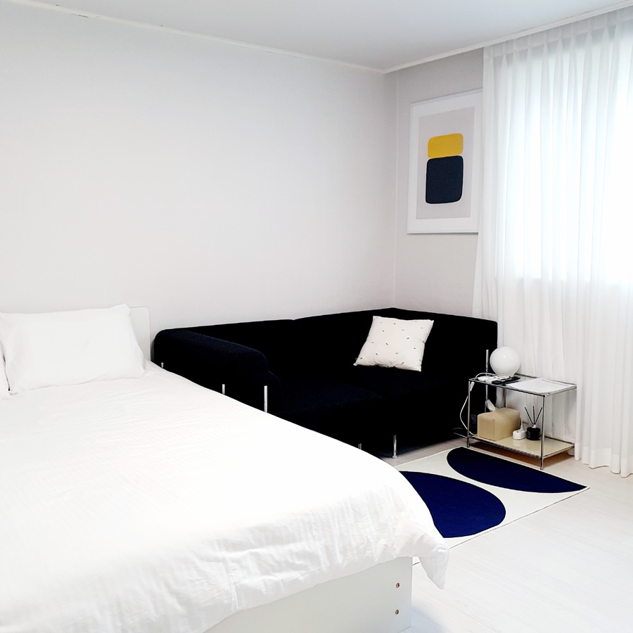 A spacious room features a white bed with neatly arranged linens. A dark-colored sofa offers comfortable seating, complemented by an accent pillow. Natural light filters through sheer white curtains, illuminating a small side table, a decorative rug, and abstract wall art.