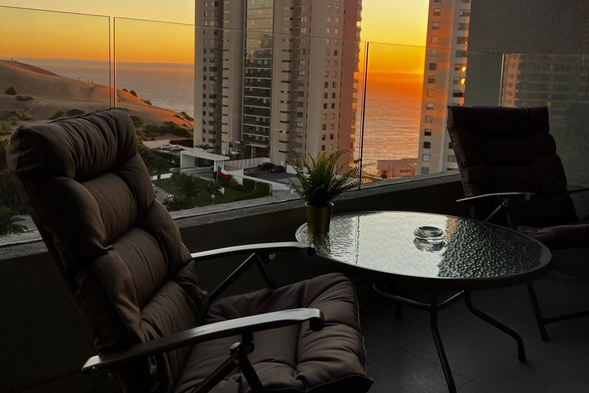 A comfortable terrace showcases two reclining chairs and a glass table. The vibrant sunset illuminates the skyline and ocean, while the nearby sand dunes create a serene backdrop. A small potted plant adds a touch of nature to the space.