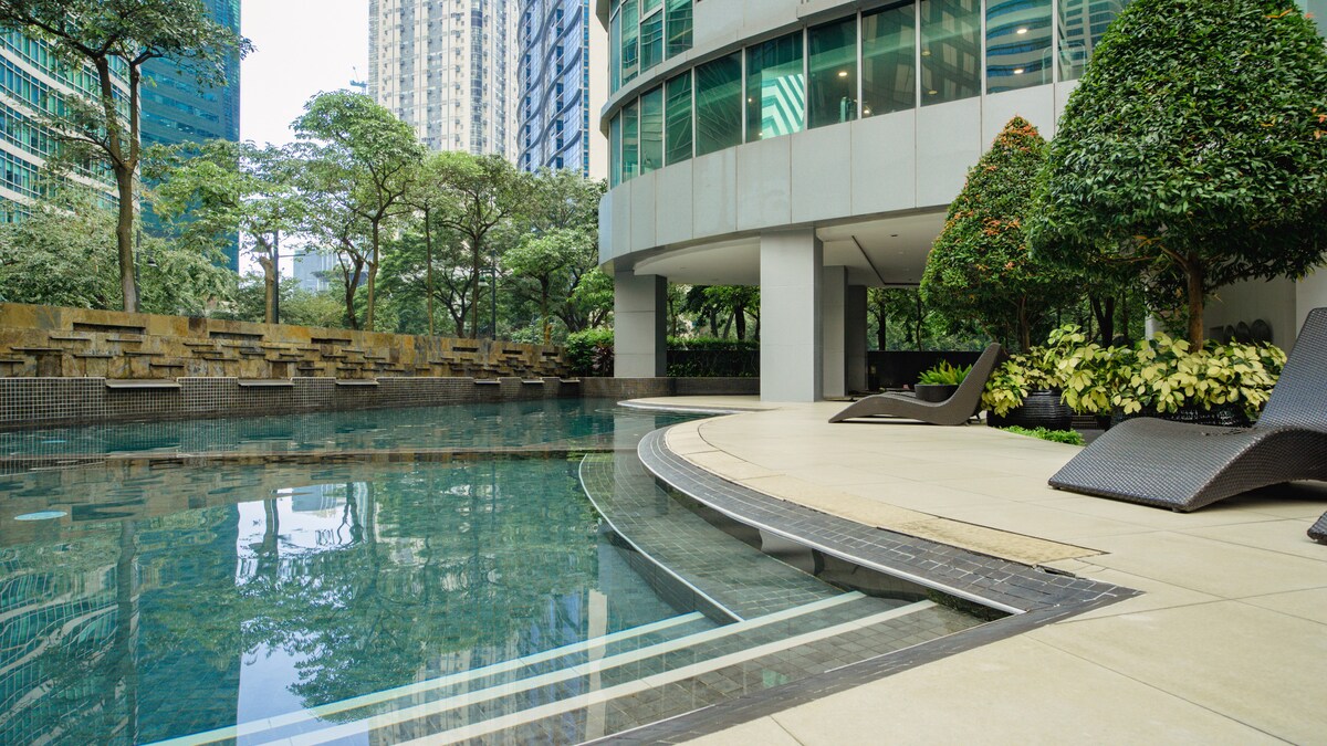The swimming pool area features a sleek design with inviting waters surrounded by lush greenery. Lounge chairs are positioned alongside the pool, offering a serene space for relaxation. The curved edges of the pool complement the modern architectural lines of the surrounding buildings.