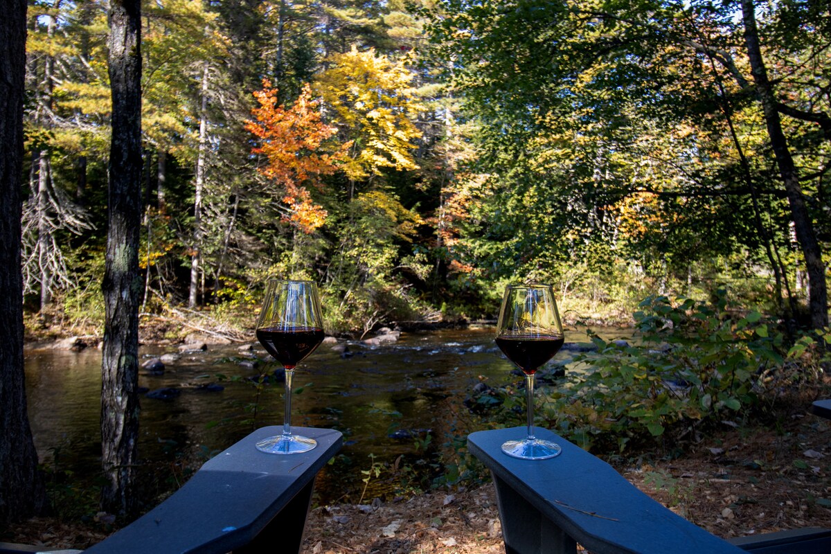Two wine glasses filled with red wine are positioned on a wooden table, overlooking a peaceful stream. Surrounding foliage displays vibrant autumn colors, while tall trees frame the scene, enhancing the natural beauty and serenity of the riverside setting.