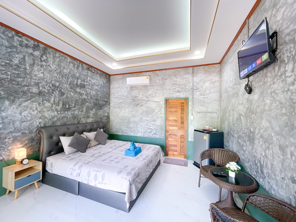 The room features a spacious bed with neutral-colored linens, flanked by two pillows. A small side table holds a lamp, while a television is mounted on the wall. Two chairs with a table are positioned near the door, and a mini fridge is visible in the corner.