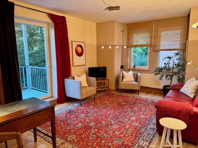 A spacious living room features a bright red rug and comfortable seating, including two armchairs and a sofa. Natural light filters through the balcony doors, illuminating the room. A small table and television are positioned for convenience, complemented by greenery from a nearby potted plant.