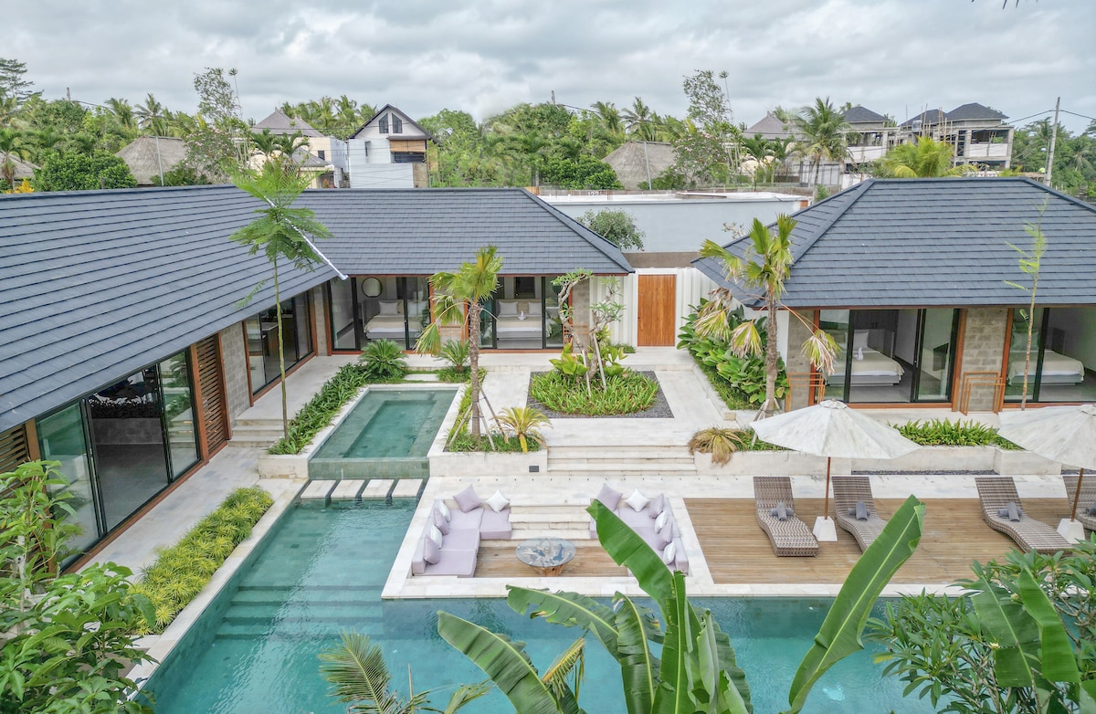 An aerial view of a spacious villa compound is displayed, featuring a large L-shaped pool surrounded by palm trees and lush greenery. Sun loungers and umbrellas are visible along the wooden deck, while modern architecture is shown in the connected living spaces.