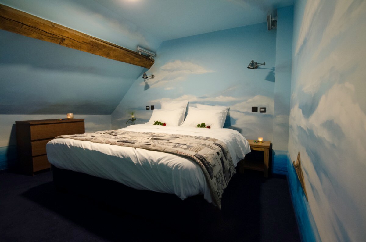 A serene bedroom features a spacious bed adorned with white linens and a decorative blanket. The walls are painted with calming skies and clouds. A wooden dresser is located nearby, with a bedside table holding candles, contributing to a cozy ambiance.