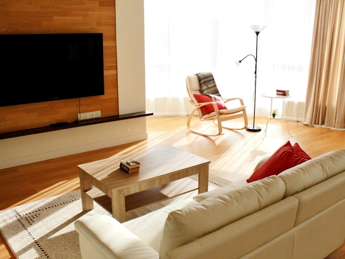 A bright living area features a large flat-screen television mounted on a wooden wall. A light-colored sofa is positioned in front of a wooden coffee table, while a cozy armchair and floor lamp provide additional seating, all bathed in natural light from large windows.