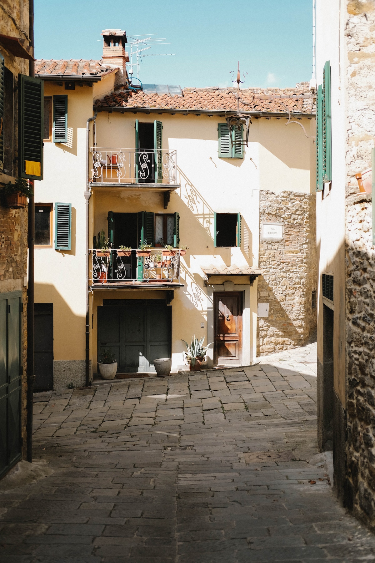 A narrow cobblestone alleyway features a charming building with a warm yellow facade. Green shutters are seen adorning the windows, while a wrought-iron balcony displays potted plants. A wooden door opens into the structure, providing a glimpse into the inviting space beyond.