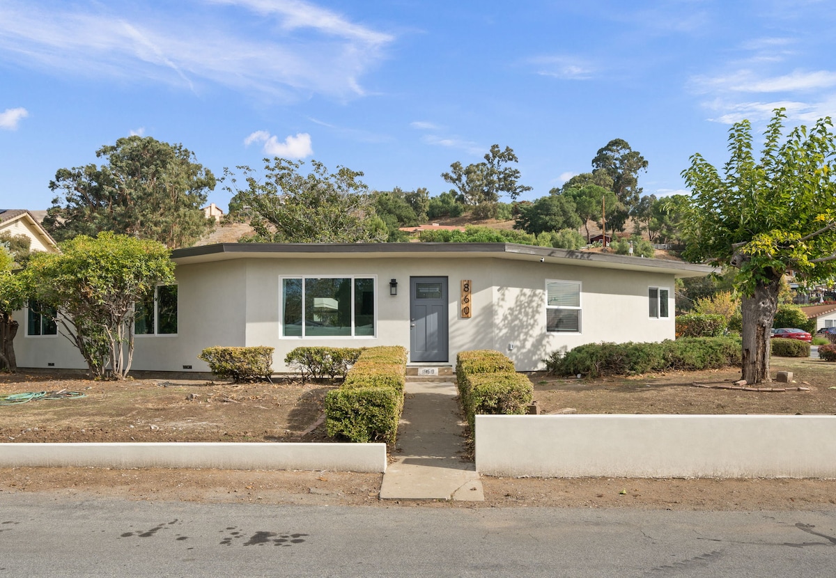 The modern exterior of a single-story home is showcased, with a rounded front and large windows allowing natural light. Lush greenery lines the entrance, and the property is bordered by a low wall along the sidewalk, creating an inviting approach.