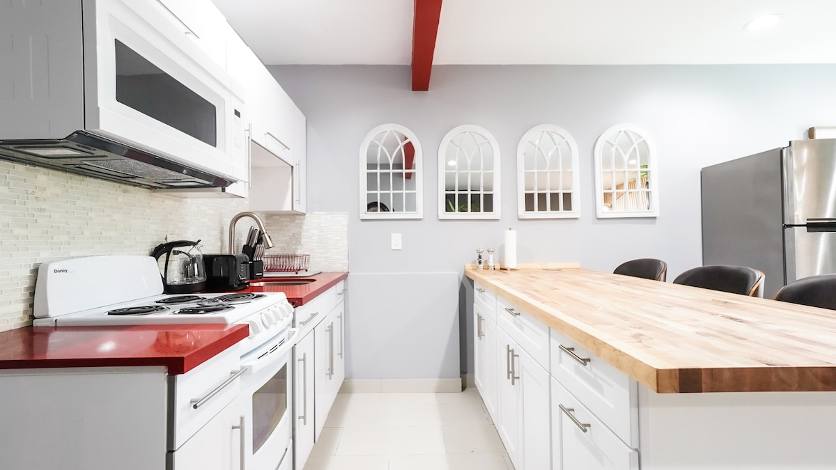 A modern kitchen features a red accent wall and a large wooden island with ample seating. Stainless steel appliances, including a microwave and refrigerator, are highlighted. Four arch-shaped windows allow natural light to enter, enhancing the bright and functional space.