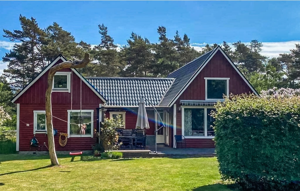 A charming red house with a sloped roof is surrounded by lush greenery. A patio area is furnished with outdoor seating and an umbrella, providing a space for relaxation. Trees and shrubs create a natural backdrop, emphasizing the property's serene outdoor setting.