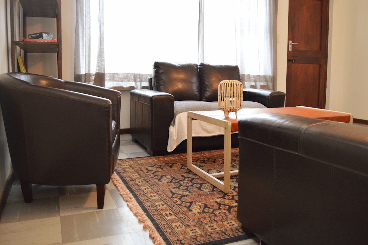 A cozy living area features dark leather sofas facing each other, placed on a patterned rug. A wooden coffee table sits centrally, with natural light filtering in through sheer curtains framing the window. A small bookshelf is positioned in the corner.