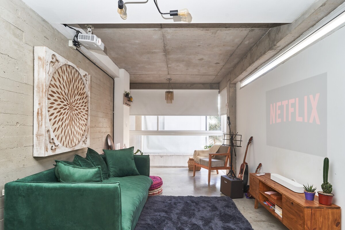 A spacious living area features a plush green sofa against a concrete wall, with a decorative wooden art piece nearby. A projection screen displaying 'NETFLIX' emphasizes the entertainment option. A wooden shelf holds a guitar and plants, enhancing the comfort of the space.