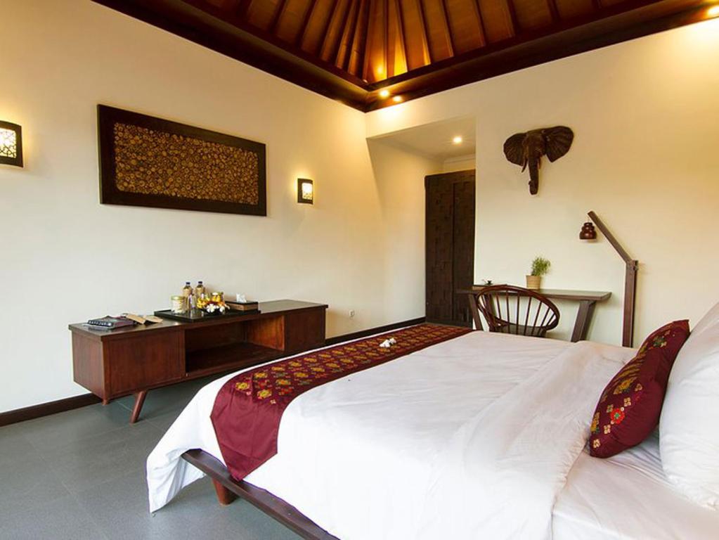 A well-appointed room features a spacious bed adorned with a decorative maroon throw. A wooden desk and chair are positioned nearby, while a console table displays beverages. Soft lighting is provided by wall sconces, and an elegant wall hanging adds a touch of character.