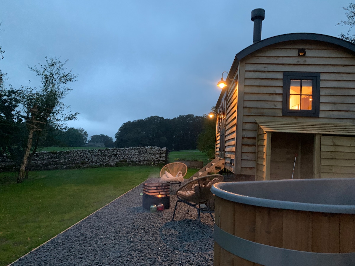 A wooden hut stands in a landscaped garden area featuring a wood-fired hot tub. Soft lighting highlights the exterior, while a fire pit surrounded by seating offers a cozy gathering spot. Scenic countryside views are visible in the background, enhancing the tranquil ambiance.