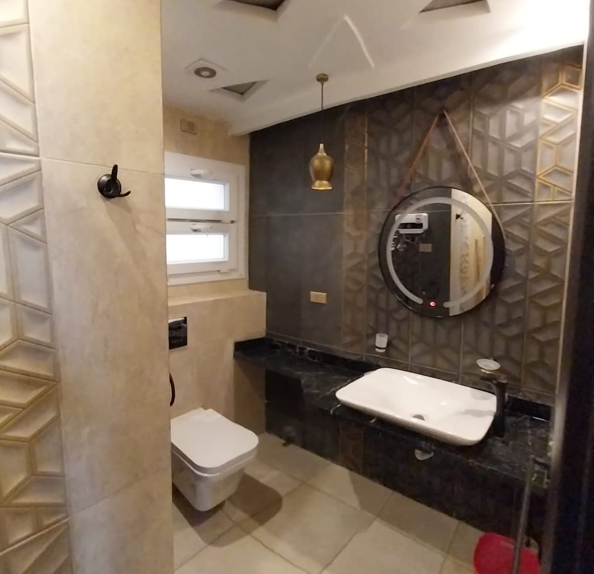 The bathroom features a combination of elegant tile designs and a modern aesthetic. A round mirror hangs above a sleek black countertop, with a white basin integrated into the surface. Natural light enters through a window, while a stylish pendant light adds illumination.