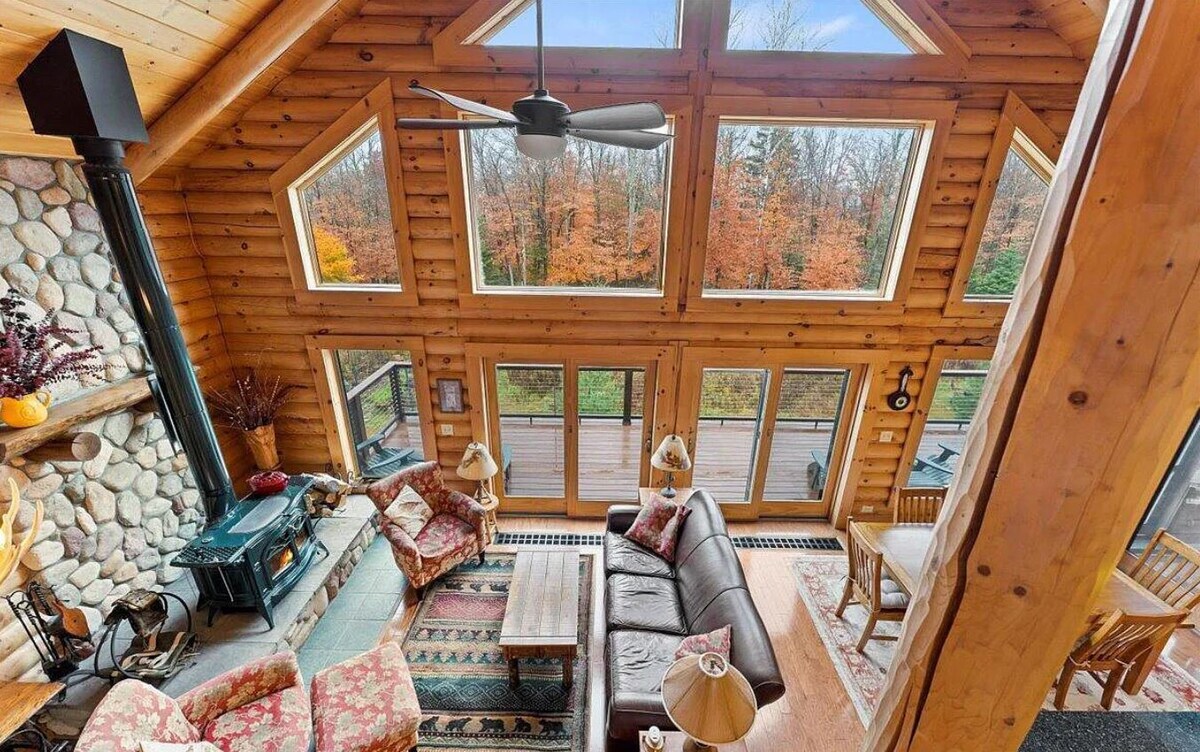 The great room features a stone fireplace and large windows that frame views of the surrounding forest. Seating options include a leather sofa and patterned chairs, with a wooden coffee table positioned at the center. A ceiling fan circulates air above, enhancing the cozy atmosphere.