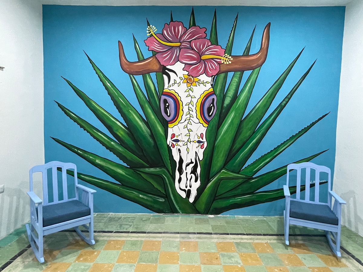 A vibrant mural featuring a decorated skull is displayed prominently on a blue wall, surrounded by large green agave leaves. Two light blue chairs with dark cushions are positioned in front of the mural, offering a comfortable seating area.