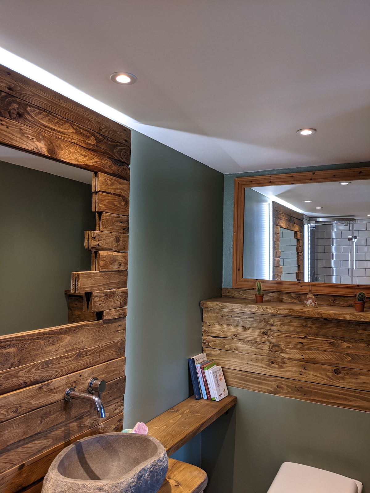 The bathroom features rustic wooden accents on the walls and mirror, complemented by modern lighting. A stone wash basin sits on a wooden shelf, providing a natural touch. The shower area is visible in the background, showcasing a sleek design with tiles.