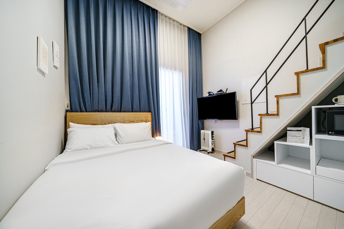 A spacious sleeping area is presented with a comfortable bed covered with crisp white linens. Natural light filters through sheer curtains, illuminating the room. A television is mounted on the wall opposite the bed, while a staircase leads to an upper level.