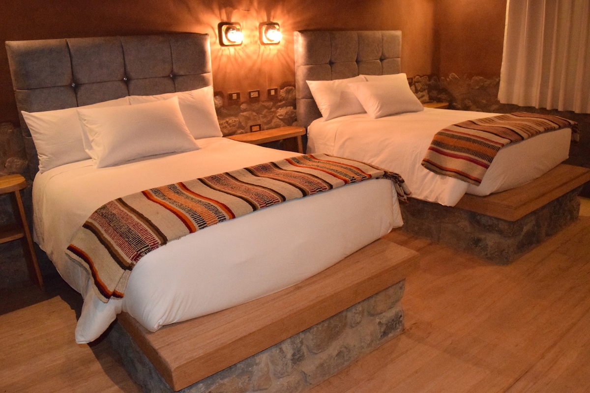 Two spacious beds are positioned in the room, each with a decorative blanket draped across the foot. Soft lighting creates a cozy ambiance, while stone accents and wooden furnishings add a rustic charm. Neutral tones dominate the space, enhancing the overall comfort.