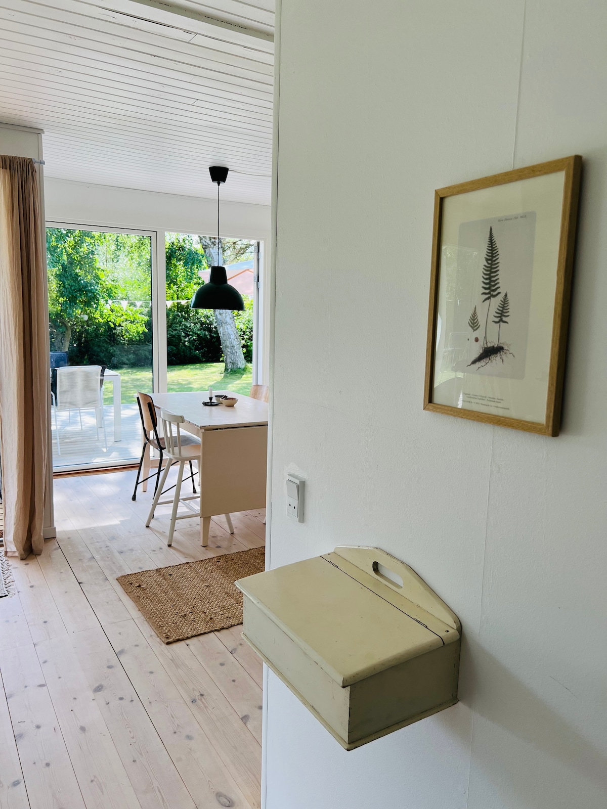 A bright entryway leads into a living space adorned with natural light from large glass doors opening to the garden. A simple dining table and chairs are present, complemented by a stylish pendant light. A small mail holder enhances the welcoming atmosphere.