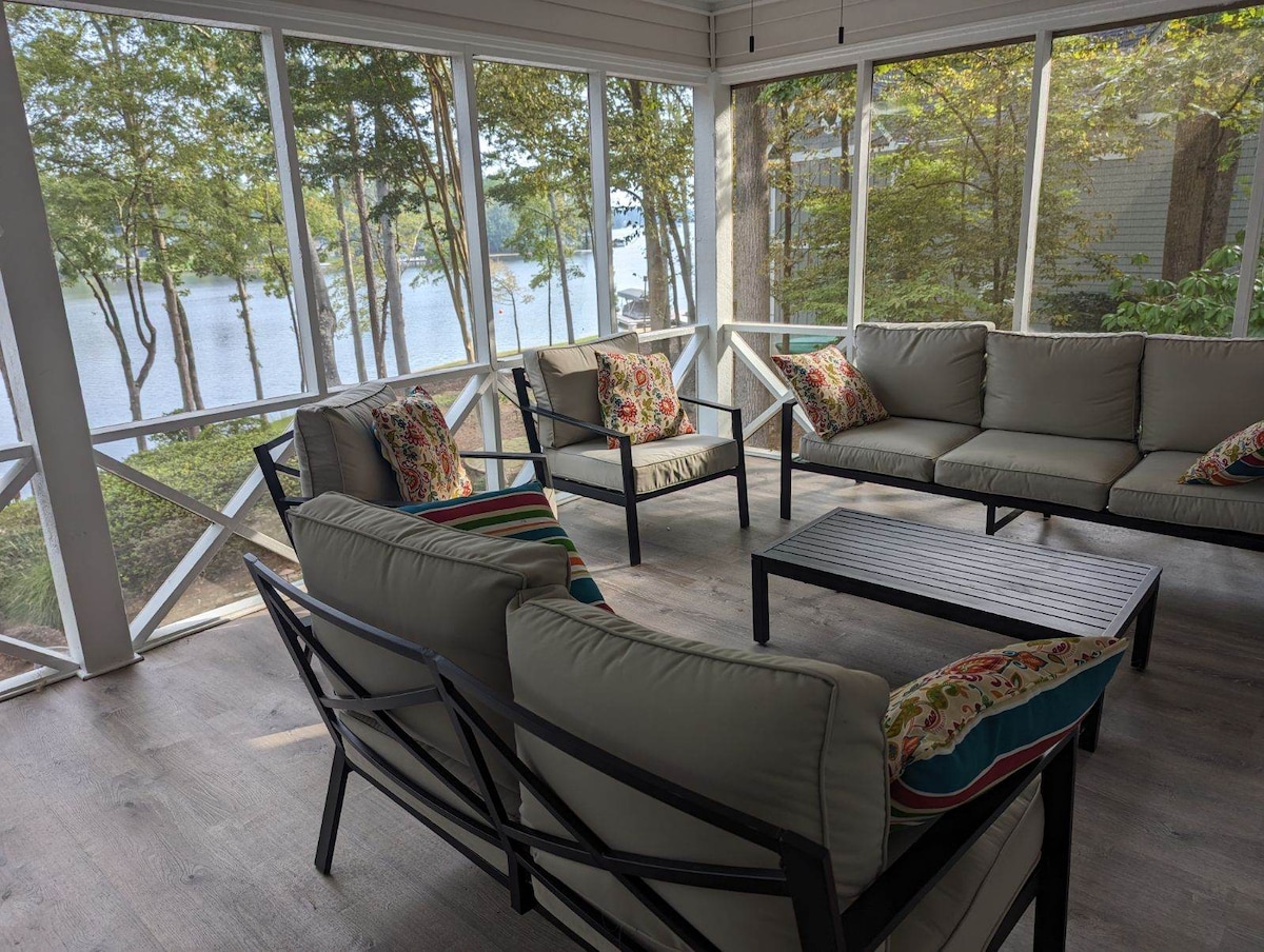 A screened porch is shown with comfortable seating arranged around a coffee table. Colorful cushions add a touch of vibrancy. Large windows provide uninterrupted views of the lake and surrounding trees, creating a serene environment for relaxation.