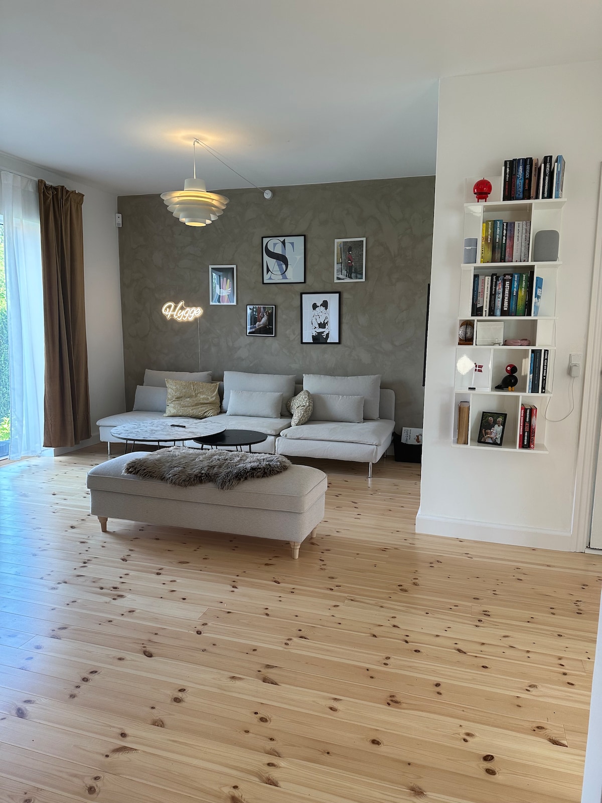 A light-filled living room features a neutral color palette with wooden flooring. A comfortable seating area includes a sofa adorned with decorative cushions. Wall art and bookshelves add character to the space. Natural light streams in through large windows, enhancing the inviting ambiance.