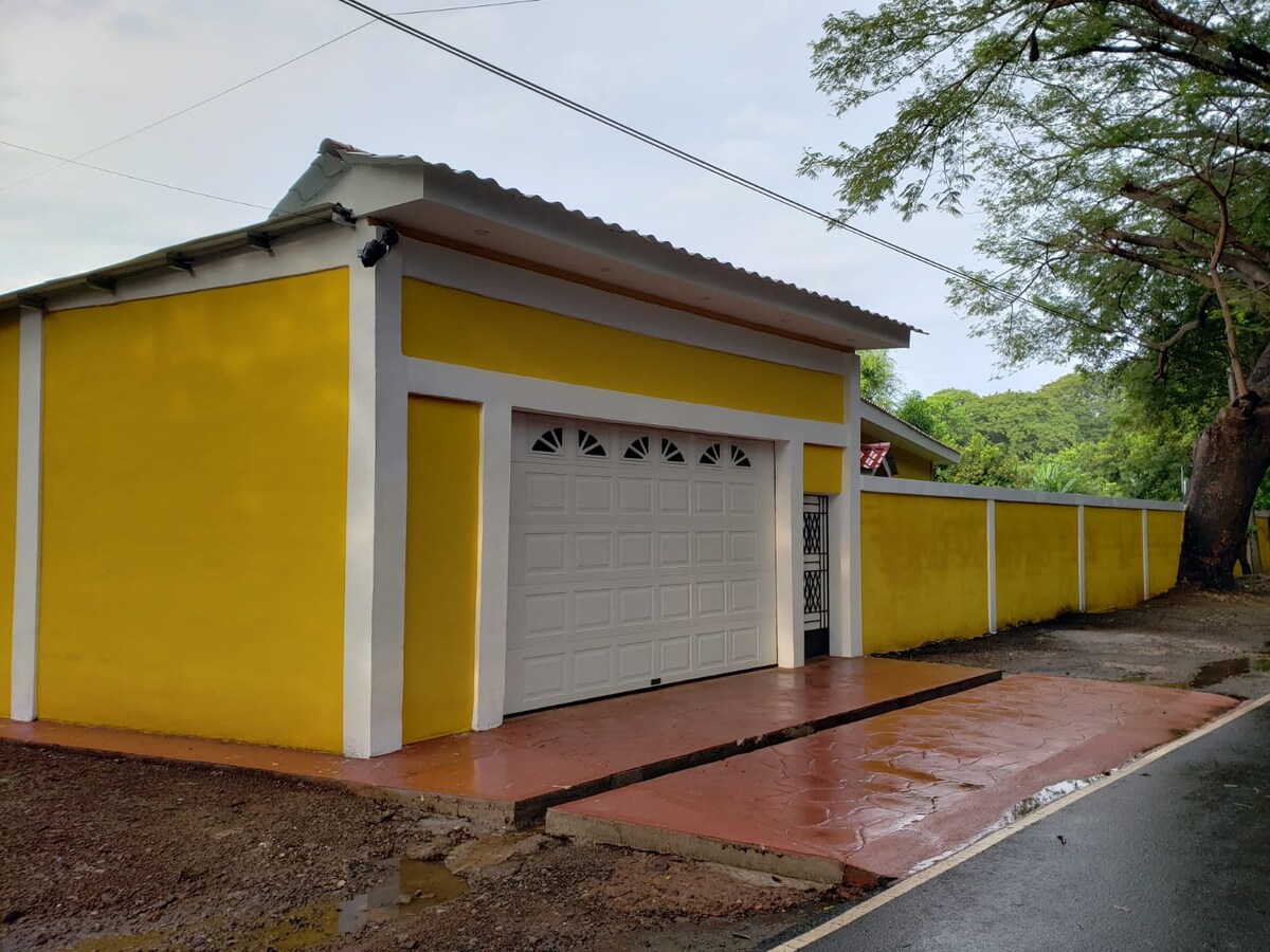 A bright yellow exterior of the house is presented, featuring a white garage door and simple architectural details. The well-maintained path leads to a gated entry, with lush greenery surrounding the property and a clear sky above.