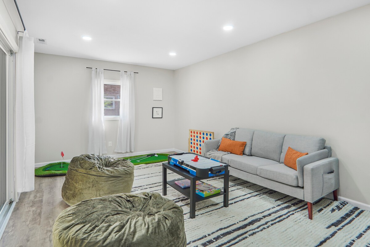 A bright game room features a cozy couch and two plush bean bags, inviting relaxation. A mini golf area and colorful board games are visible. Natural light fills the space through a window framed by sheer curtains, adding to the room's playful atmosphere.