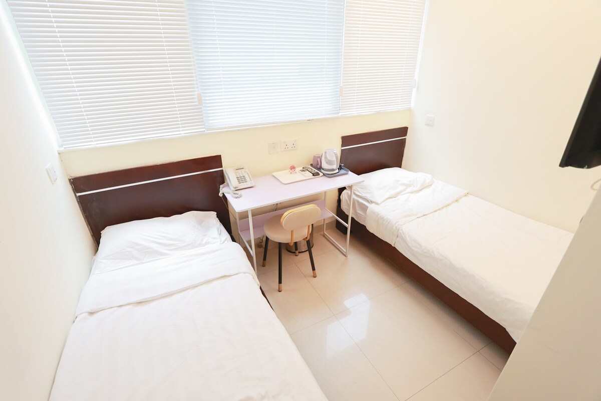 A room featuring two spacious beds, both dressed in white linens, is illuminated by natural light from large windows. A simple desk with a chair is positioned between the beds, and a flat-screen TV is mounted on the wall, providing essential comfort for guests.