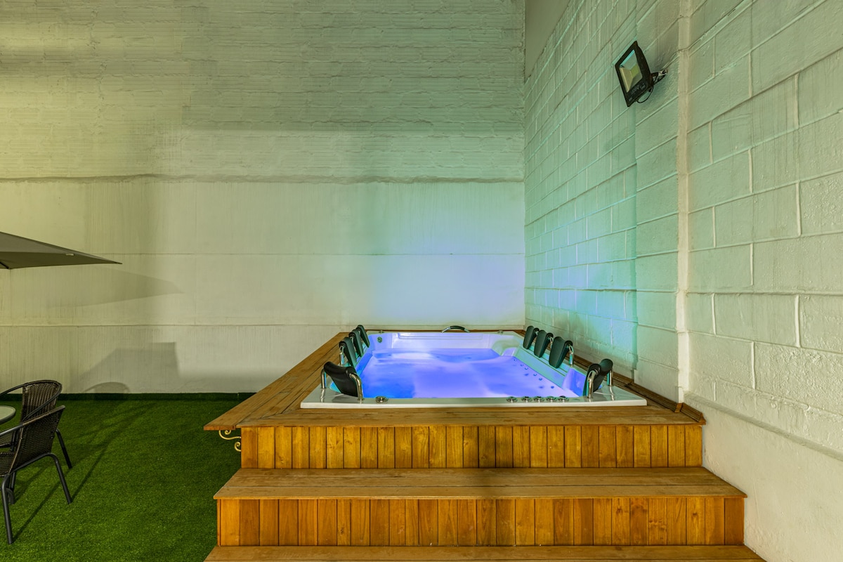 A spacious hot tub is set against a white wall, surrounded by wooden decking. Soft lighting enhances the ambiance, while artificial grass adds a touch of greenery. A few chairs are visible in the area, providing additional seating for relaxation.