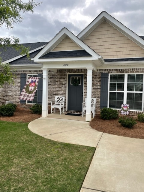 The home's entrance features a welcoming porch with two white chairs, a decorative wreath hanging on the door, and well-maintained landscaping. Concrete pathways lead up to the front door, framed by brick and siding in soft earth tones.