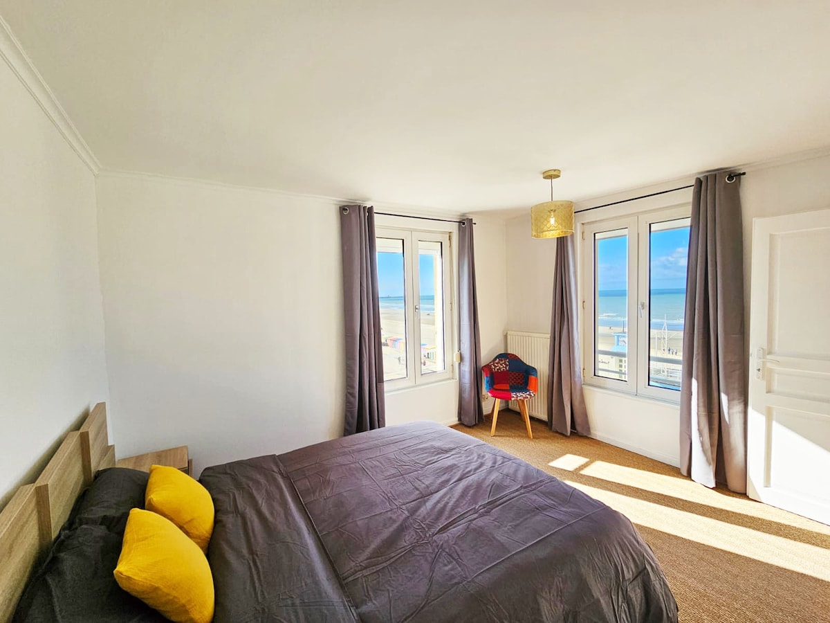 A bright bedroom features large windows offering a view of the sea. A spacious bed with dark bedding is complemented by two yellow pillows. Sheer curtains frame the windows, allowing natural light to fill the room, which includes a modern light fixture and a colorful chair.