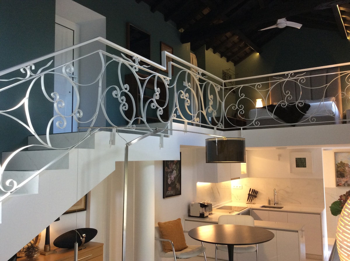 An elegant loft space is highlighted by a stylish metal railing along a curved staircase. The open kitchen features modern appliances and a round dining table, while the upper level showcases a cozy sleeping area with soft lighting and wooden beams.