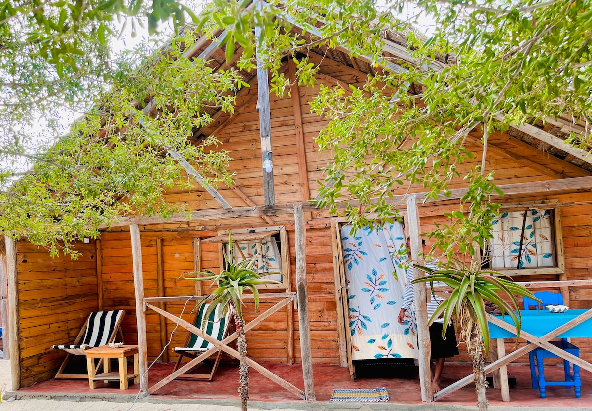 A rustic wooden bungalow is framed by lush green foliage. Two striped chairs provide seating on the porch, while a vibrant blue table is positioned nearby. Floral-patterned curtains are visible inside through open doors, adding an inviting touch to the exterior.