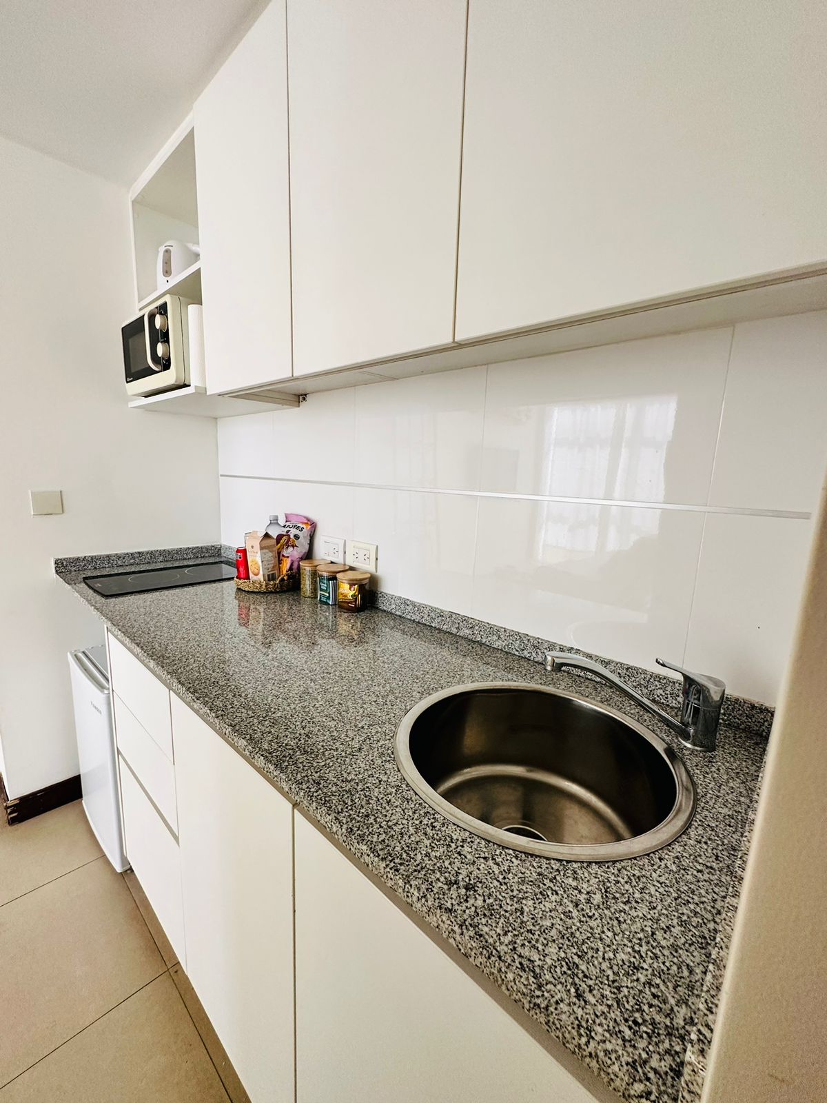 A compact kitchen area is equipped with white cabinetry and a granite countertop. A stainless steel sink is positioned beside a small refrigerator and a microwave. Pantry items are arranged neatly on the counter, providing accessibility for meal preparation.