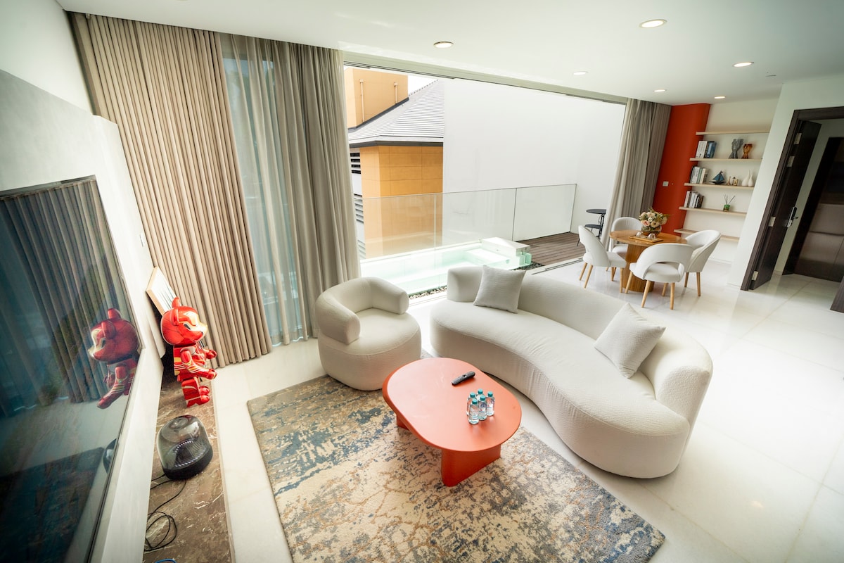 A modern living room features a plush, light-colored sofa set paired with a round coffee table. Large glass doors provide natural light and views of the outdoor balcony. Soft, neutral tones are complemented by a decorative rug and stylish artwork on the wall.