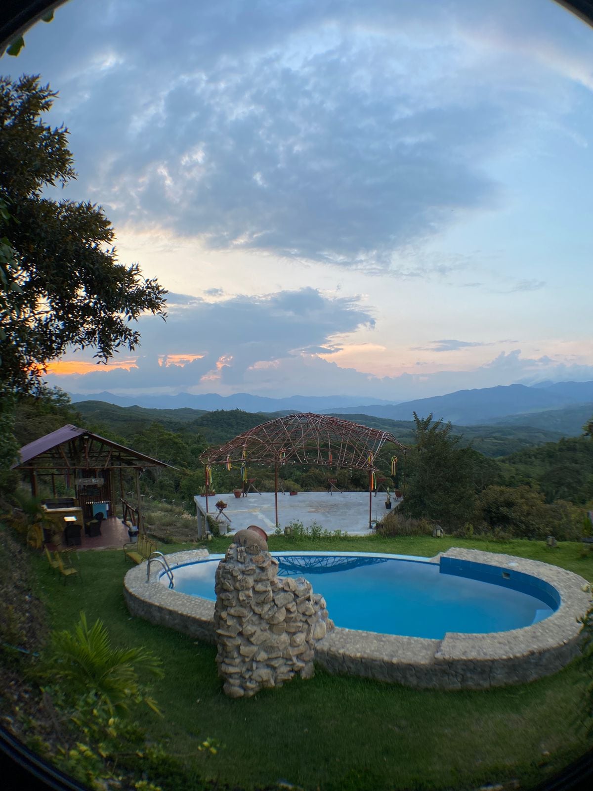 An outdoor area features a circular pool surrounded by natural stone, with a distant mountain range under a soft sunset sky. A gazebo structure stands nearby, complemented by a rustic building, lush greenery, and scenic views of the hills.