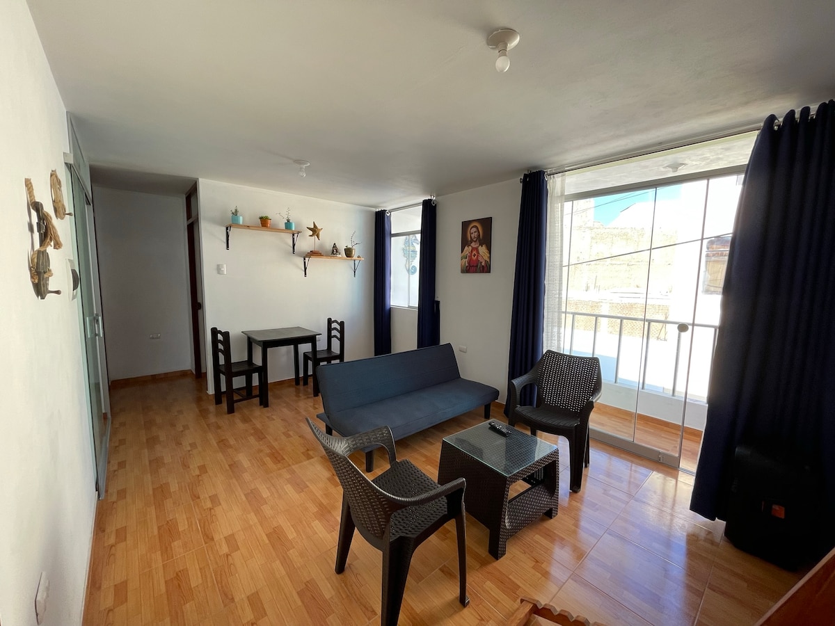 A common area is featured with light-colored wooden flooring. A dark sofa and two chairs surround a glass coffee table. A small dining table is located in the background, accompanied by a few decorative items on the walls and a large window allowing natural light.