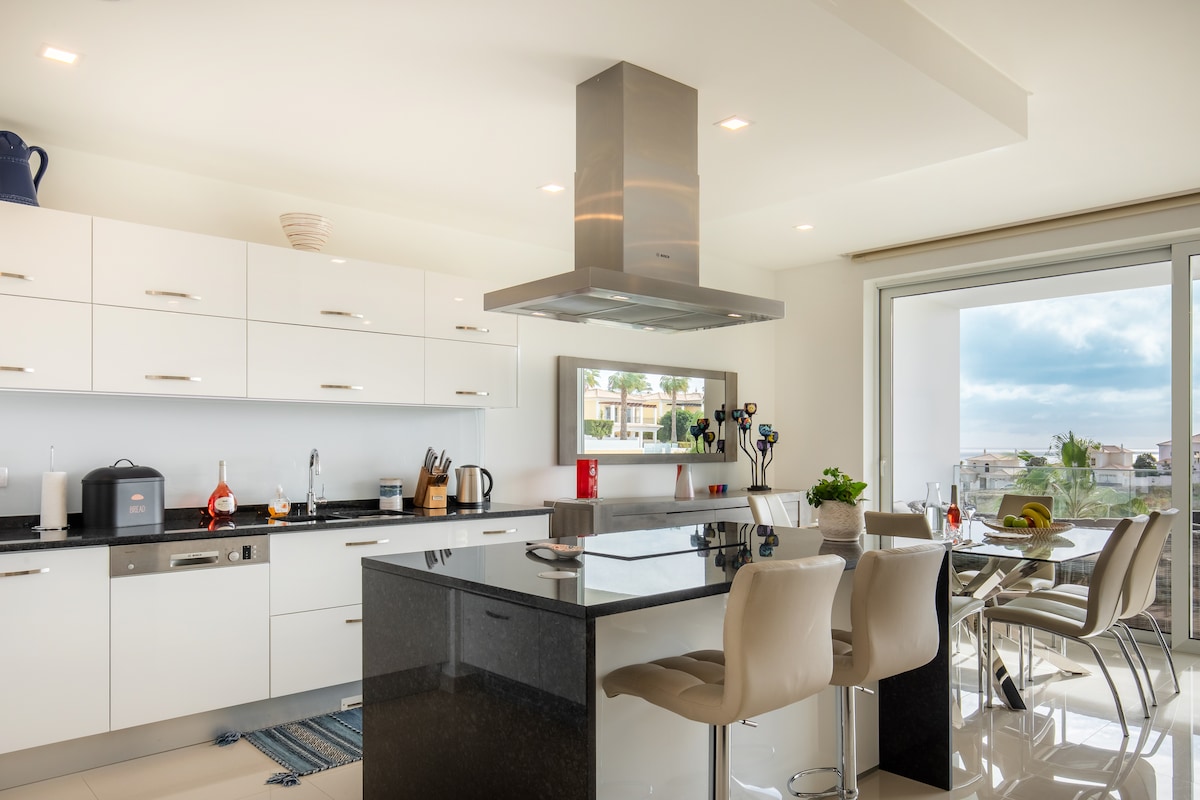 A modern kitchen features sleek white cabinetry and a dark countertop. High-end appliances are neatly arranged, with a large island offering seating. Natural light floods the area through a wide window, providing a view of the outdoors. Decorative elements and utensils are present on display.