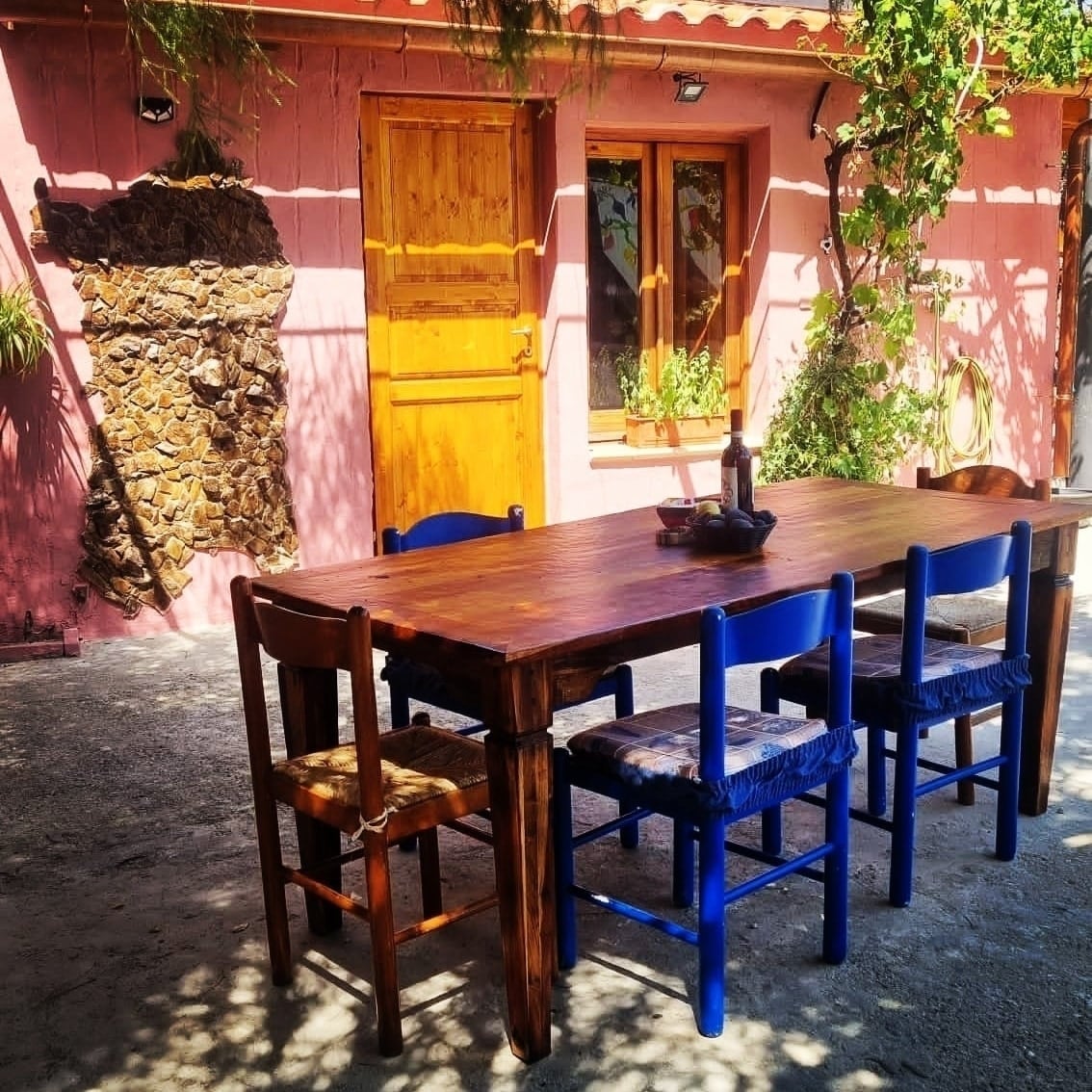 A rustic wooden dining table, surrounded by six chairs in natural wood and blue, is positioned outside against a pink wall adorned with a stone accent. A bottle of wine and glasses are set on the table, inviting outdoor dining under dappled sunlight.