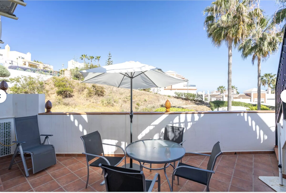 A shaded outdoor terrace features a round table surrounded by four black chairs. A large umbrella provides shade, while palm trees and a hillside landscape are visible in the background under a clear blue sky.