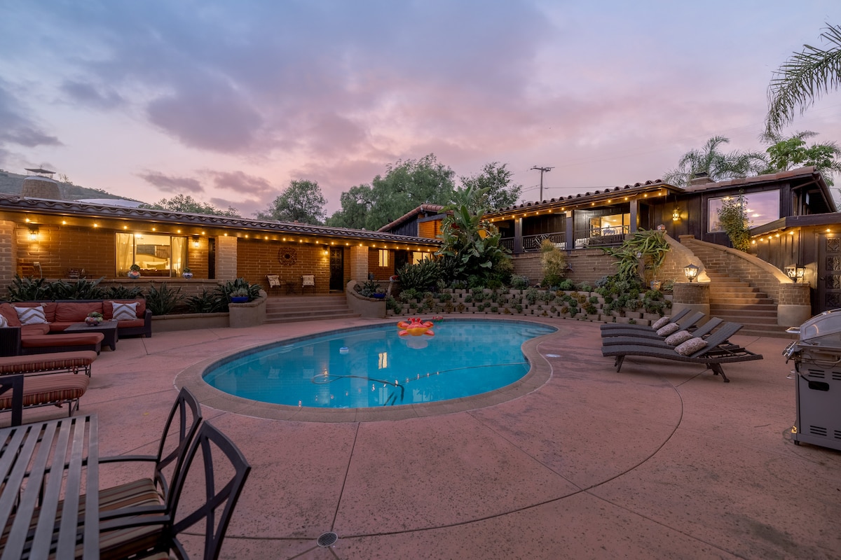 A serene outdoor space is highlighted, featuring a salt-water pool surrounded by lounge chairs. Warm lighting casts a soft glow on the brick walls. Steps lead to a higher level, adorned with lush plants, creating a welcoming atmosphere for relaxation.