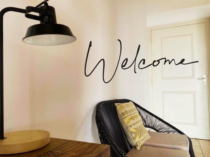 A comfortable seating area is shown, featuring a light fixture with a black shade positioned near a sleek wooden table. A welcoming message is artistically displayed on the wall, complementing the cozy chair adorned with a decorative pillow.