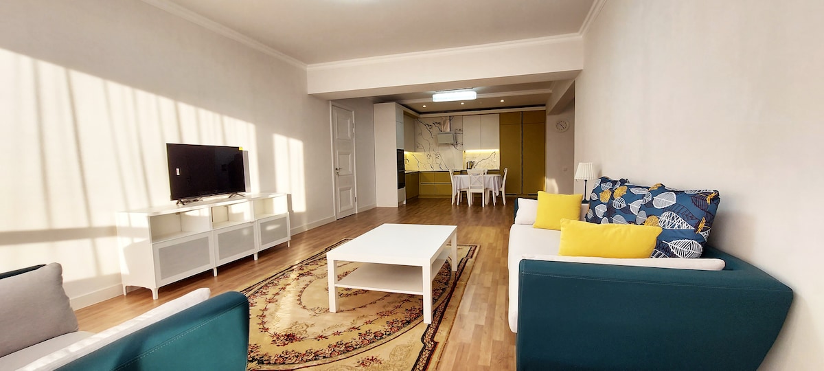 A bright living area features a neutral-toned rug at its center, surrounded by two modern sofas with cushions in blue and yellow. A low white coffee table sits in front of a console with a television. Natural light streams in through large windows, illuminating the open space.