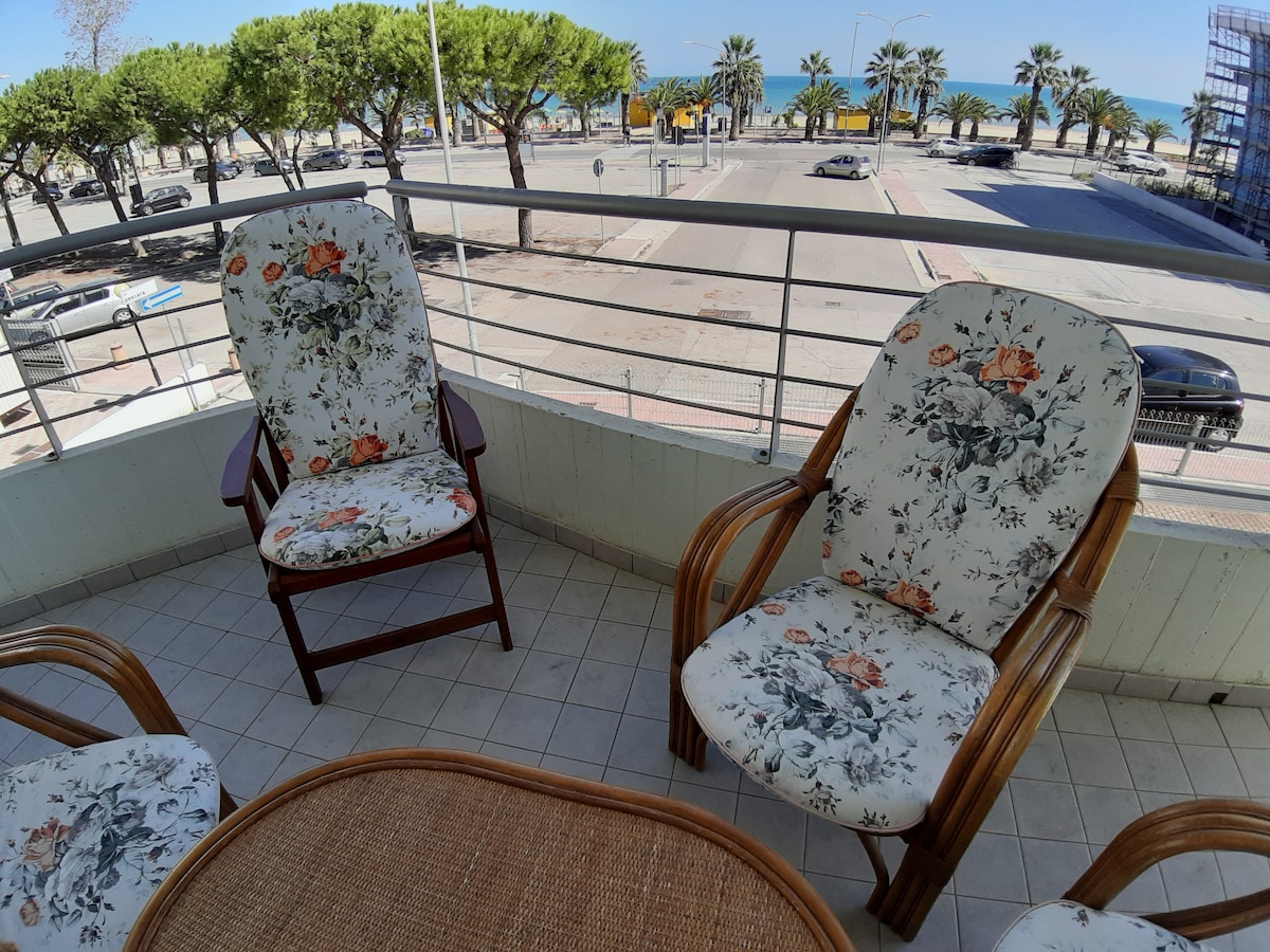 A balcony seating area features two wooden chairs with floral-patterned cushions. A small round table sits between them on a tiled floor, while views of palm trees and the ocean are visible in the background.