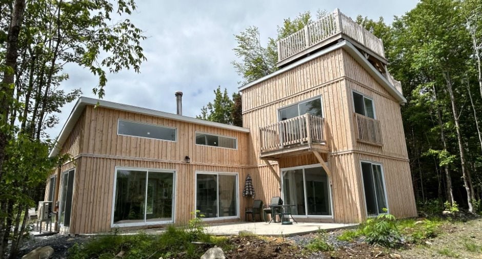 A modern, wooden two-story house features large glass doors and a spacious balcony that extends from the upper level. The surrounding greenery offers a serene backdrop. The exterior showcases natural wood paneling, emphasizing the home's connection to nature.