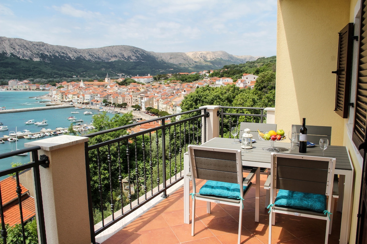 A balcony table set for dining overlooks a scenic view of a coastal town, with a table laid with fruit and drink. The surrounding landscape features lush greenery and mountainous terrain, while the water reflects blue hues and is dotted with boats.